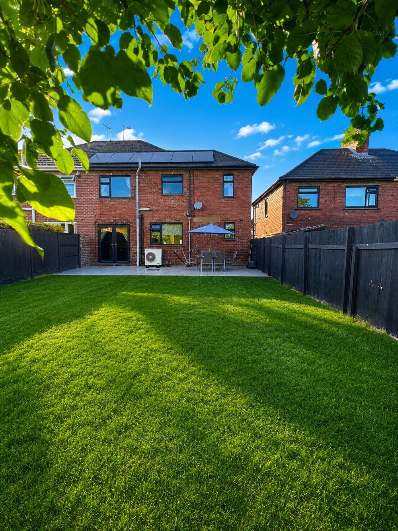 A two-story brick house with a well-maintained lawn, a patio with outdoor furniture, and a black fence surrounding the property.