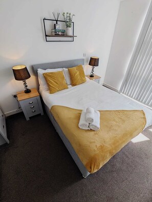 Ein ordentlich bezogenes Bett mit weißer und goldener Bettdecke, zwei Nachttische mit Lampen sowie ein an der Wand befestigtes Regal mit Pflanze und Büchern.
