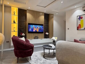 Een moderne woonkamer met een flatscreen televisie aan een houten wand, een rode fauteuil, een ronde salontafel met boeken en een vaas met bloemen.