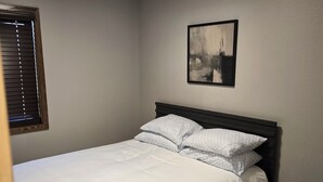 Un dormitorio con cama, mesita de noche, ventana con persianas y un cuadro colgado en la pared.
