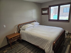 A bedroom with a wooden bed, a nightstand, a wall-mounted air conditioner, and a window with blinds.