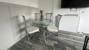 A dining area with a glass table surrounded by four chairs, a vase with dried flowers, and a mounted television on the wall.