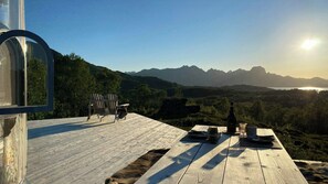 A wooden deck with outdoor seating, a bottle, and glasses, overlooking a mountainous landscape.