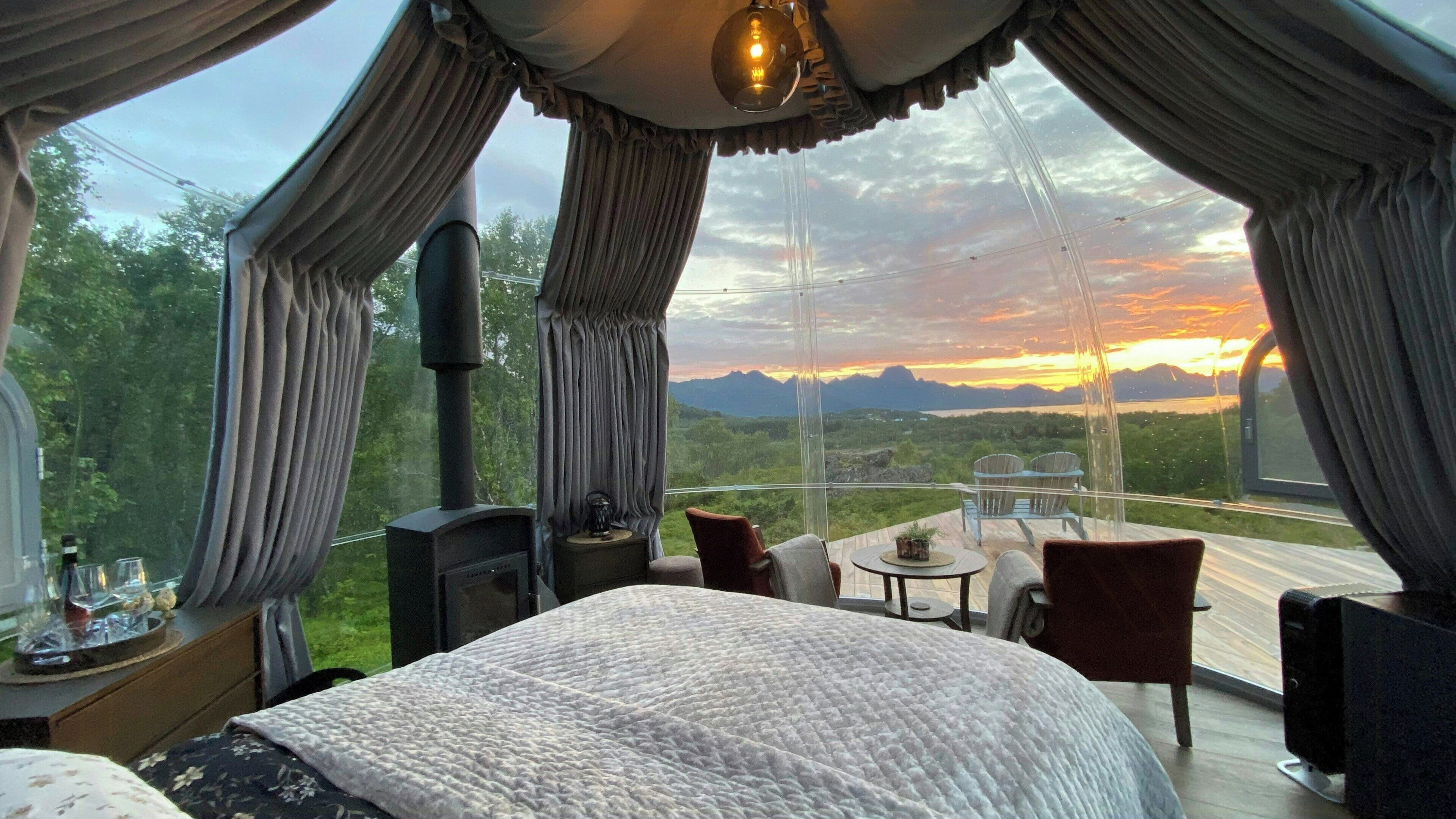 Ein Schlafzimmer mit einem großen Bett, einem kleinen Tisch, zwei Stühlen und Blick auf einen Sonnenuntergang über Bergen.