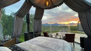 A bedroom with a large bed, a small table, two chairs, and a view of a sunset over mountains. - Glass Dome with 360° Fjord & Mountain Views (Sortland)
