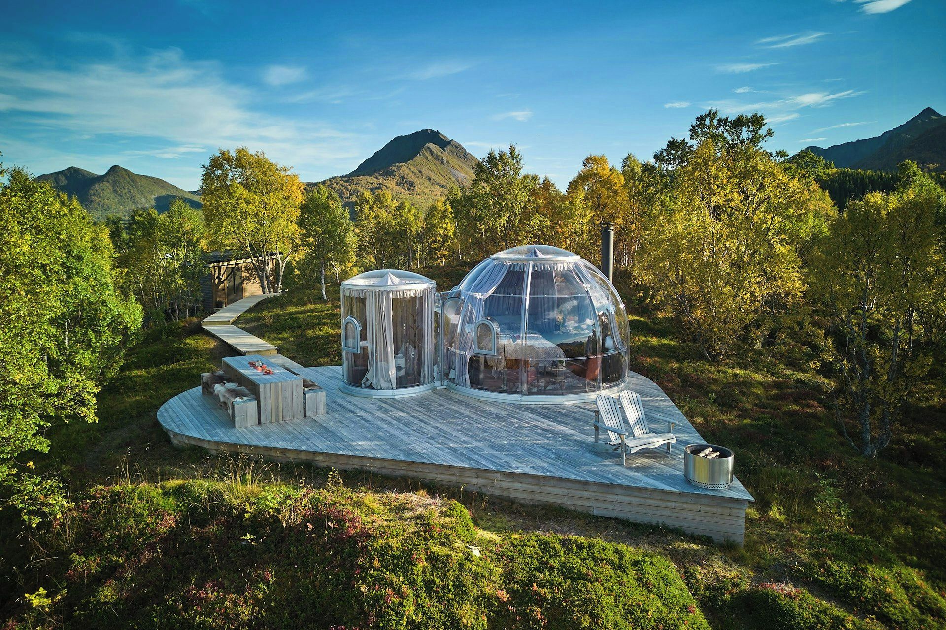 A transparent dome structure with a wooden deck, surrounded by a forested landscape and mountains.