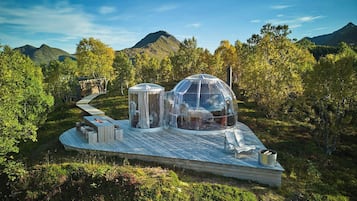 A transparent dome structure with a wooden deck, surrounded by a forested landscape and mountains.