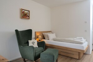 Ein schlicht eingerichtetes Schlafzimmer mit einem Einzelbett, einem grünen Sessel, einem kleinen Tisch mit Lampe und einem gerahmten Bild an der Wand.