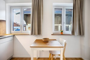 Eine Küche mit Holztisch und weißem Stuhl, einem Fenster mit Vorhängen und Blick auf Gebäude.