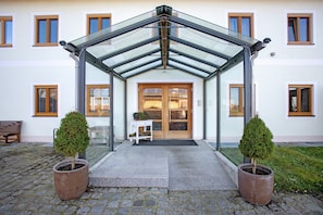 Ein Geb äudeeingang mit Vordach aus Glas, einer Holztür und zwei Topfpflanzen.