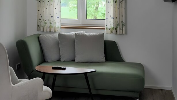 A green sofa with two pillows, a small round table, and a white chair in a room with a window and curtains. - Apartment 5 – Well-Equipped Kitchen, Spacious Outdoor Area, and Beautiful Nature Views (Kirchseeon)
