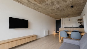 A modern living space with a flat-screen TV mounted on the wall, a wooden bench, a dining area with a table and chairs, and a kitchen area with a countertop and cabinets.