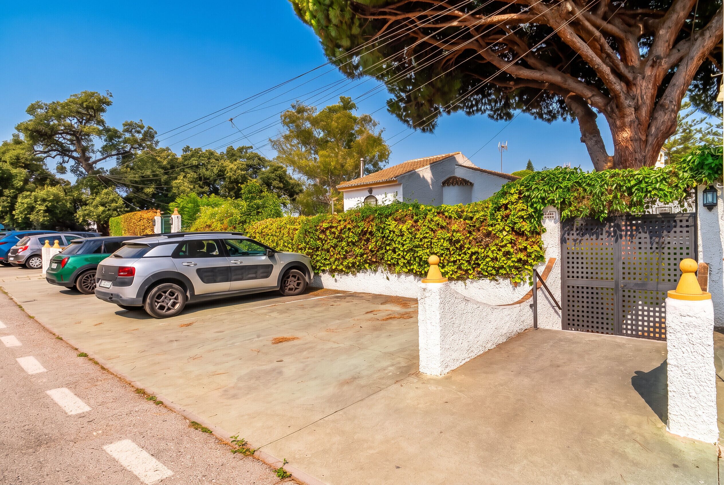 Une rue résidentielle bordée de voitures garées, une maison blanche avec un toit en tuiles et un grand arbre qui offre de l’ombre.