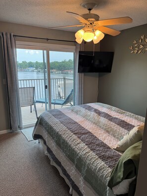 Ein Schlafzimmer mit einem großen Bett, einem Deckenventilator, einem an der Wand befestigten Fernseher und Blick auf ein Gewässer durch eine Schiebetür.