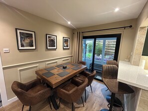 A dining area with a wooden table, brown upholstered chairs, framed pictures on the wall, and a view through a glass door to an outdoor garden. - Newly renovated cosy coastal retreat within walking distance of village pub! (Kippford)