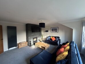 A living room with a fireplace, a black sofa, a wooden coffee table, and a black TV mounted on the wall. - Newly renovated cosy coastal retreat within walking distance of village pub! (Kippford)