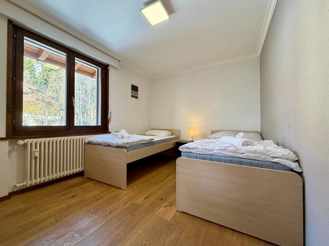 A bedroom with two single beds, a radiator, and a window with a view of trees.
