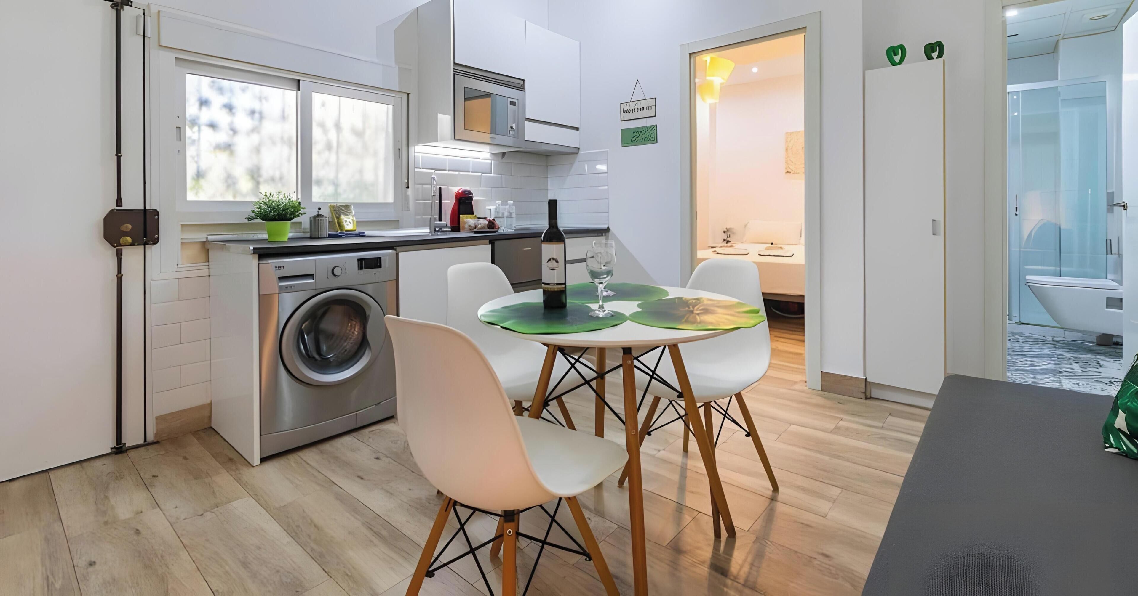 A compact kitchen with a washing machine, a dining area with a table and chairs, and a view into a bathroom.
