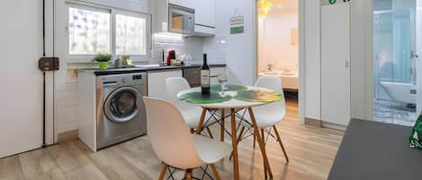 A compact kitchen with a washing machine, a dining area with a table and chairs, and a view into a bathroom.