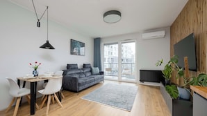 Un salon moderne avec un canapé gris, une table à manger et des chaises, un tapis gris et une grande fenêtre donnant sur des immeubles.