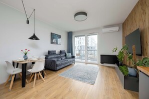 A modern living room with a grey sofa, dining table, and chairs, a grey rug, and a large window with a view of buildings. - Spacious Flat with a Balcony | Parking | Air-conditioning (Warszawa)