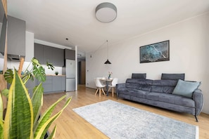 A modern living space with a grey sofa, a dining table with chairs, a kitchen area, and a large window. - Spacious Flat with a Balcony | Parking | Air-conditioning (Warszawa)