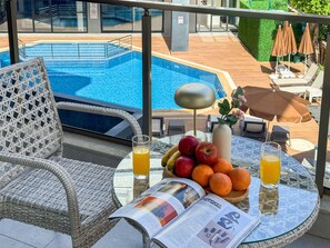 Ein Balkon mit Korbstühlen, einem Glastisch mit Früchten, Getränken und einer Zeitschrift, mit Blick auf ein Schwimmbad.