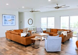 A spacious living room with two sofas, a coffee table, and a chair. There are ceiling fans, a clock, and large windows.