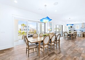 A spacious dining area with a long wooden table and chairs, large windows offering a view of the cityscape, and a modern kitchen in the background.