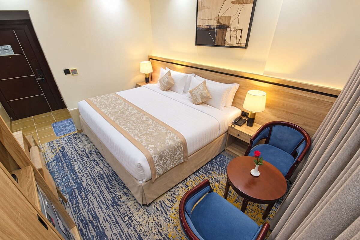 A hotel room with a bed, bedside lamps, a chair, a small table, and a painting on the wall.