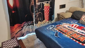 Un dormitorio con una cama cubierta por un edredón azul con diseño cinematográfico, un cartel grande en la pared y una ventana con persianas.