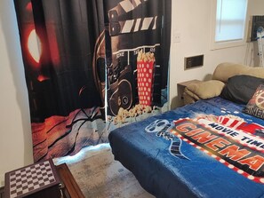 Een slaapkamer met een bed belegd met een blauwe deken met een filmthema, een groot poster aan de muur en een raam met zonwering.