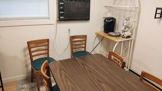 Un comedor con una mesa y sillas de madera, una máquina de café y un estante metálico blanco con hervidor y tazas.