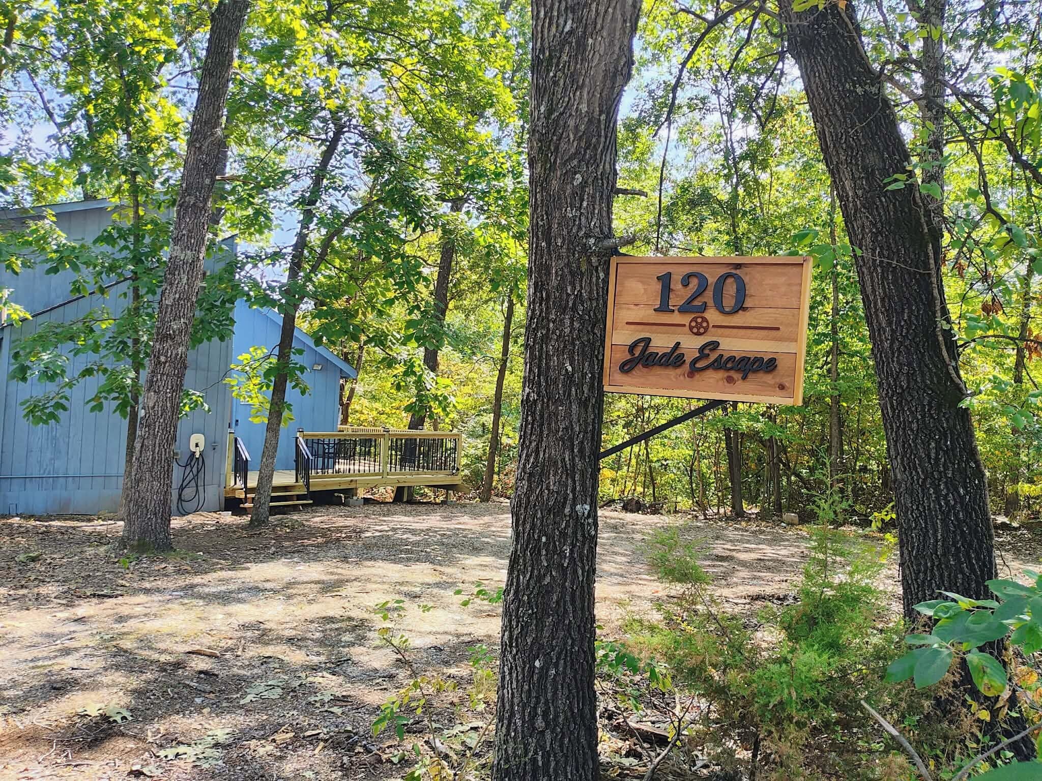 A wooden sign with the number 120 and the text 'Jade Escape' in a forested area with trees and a blue building in the background.