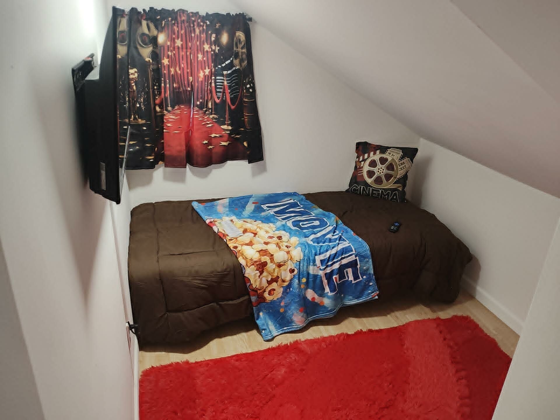 Una habitación pequeña con cama individual, alfombra roja, manta azul con diseño de palomitas y almohada con temática de cine.
