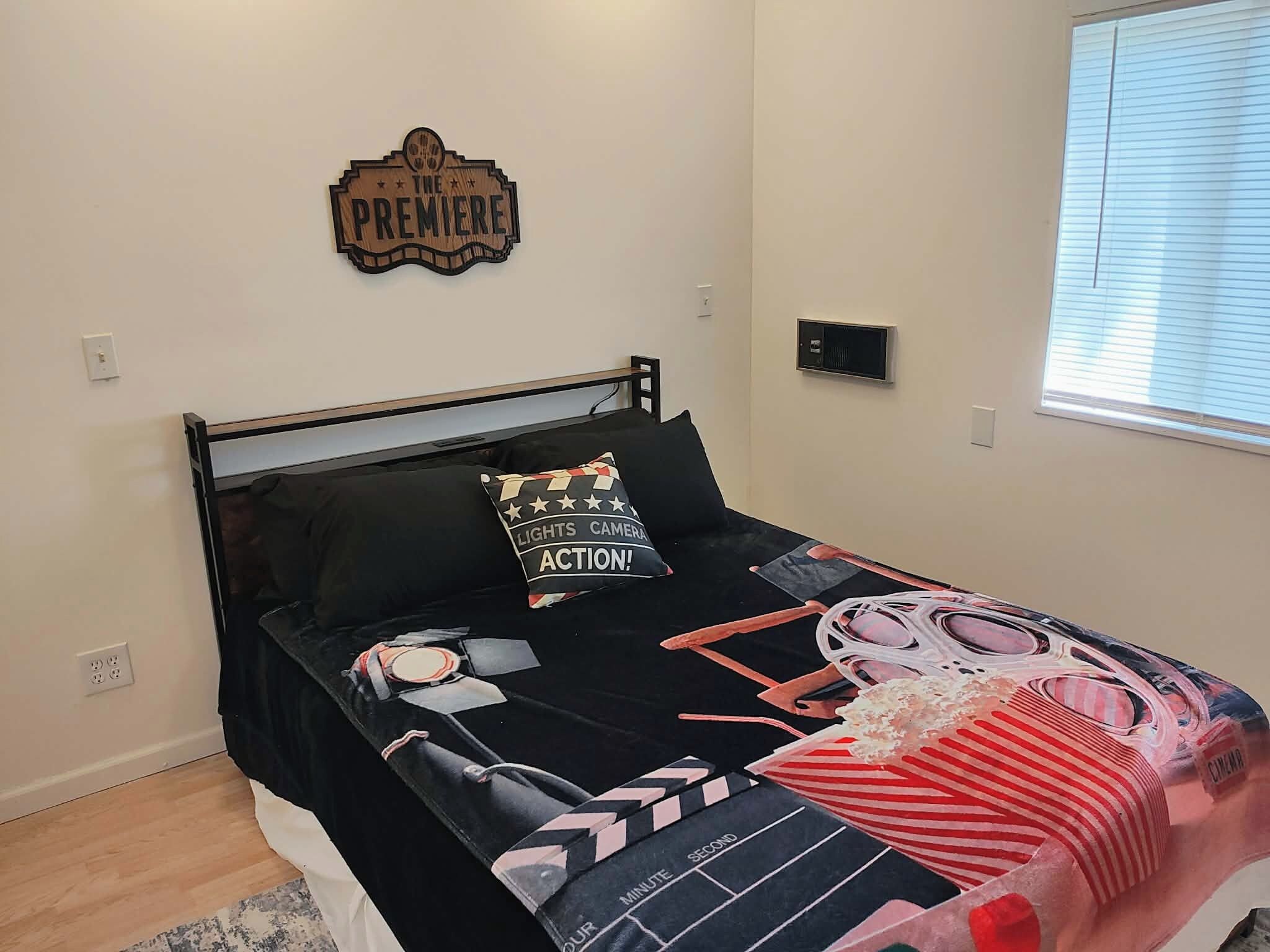 A bedroom with a bed, a pillow with a camera design, a wall clock, and a window with blinds.