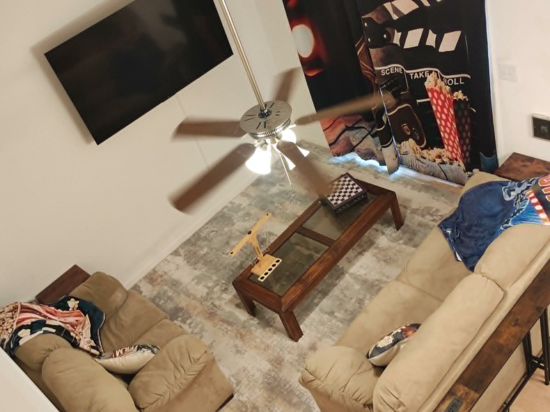 A living room with a ceiling fan, a flat-screen TV mounted on the wall, a coffee table, and two sofas.