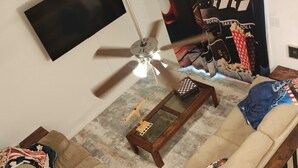 Una sala de estar con ventilador de techo, un televisor de pantalla plana montado en la pared, una mesa de centro y dos sofás.