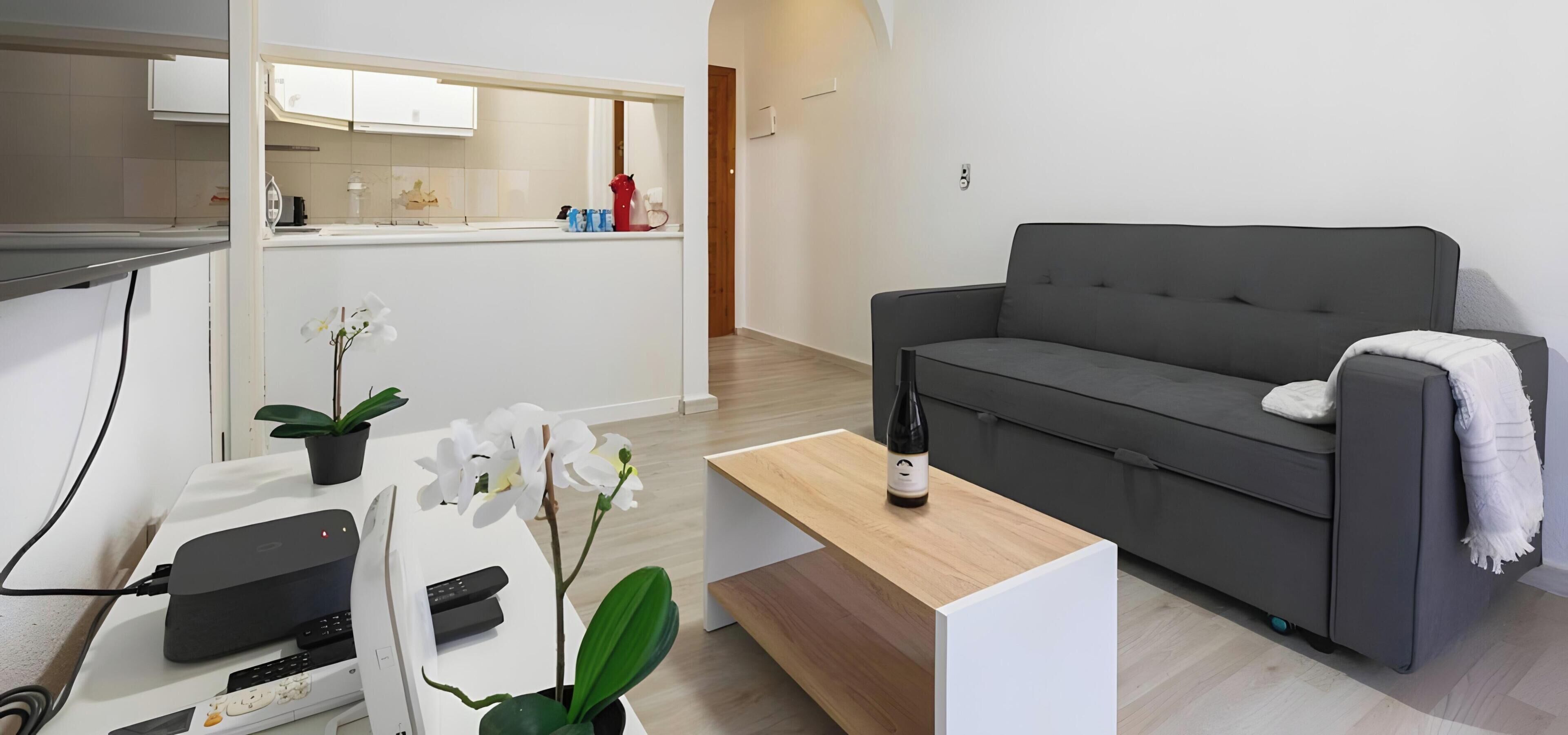 Una sala de estar moderna con un sofá gris, una mesa de centro de madera y una cocina al fondo.