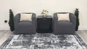 Två grå fåtöljer med vita kuddar, ett svart sidobord med en vas med blommor och en svart högtalarenhet.