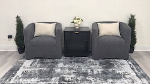Zwei graue Sessel mit weißen Kissen, ein schwarzer Beistelltisch mit Blumen in einer Vase und eine schwarze Lautsprecherbox.