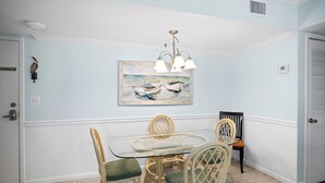 Et spiseområde med et glasbord, fletstole, et maleri af både og en loftlampe.