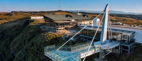 A modern building with a glass bridge and a metal framework, surrounded by a hilly landscape.