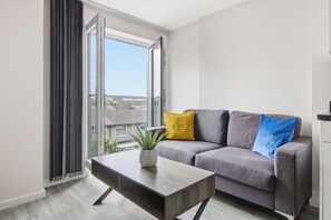 A modern living room with a grey sofa, a wooden coffee table, and a view of a residential area through large windows. - Modern 2 Bedroom Flat l Manchester city l Sleeps 5 ! (Manchester)