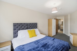 A bedroom with a tufted headboard, a bed with white and blue bedding, a yellow pillow, a wooden nightstand, and a wardrobe. - Modern 2 Bedroom Flat l Manchester city l Sleeps 5 ! (Manchester)
