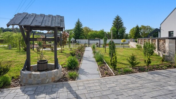 En have med et træpergola, en stenildsted, en belagt sti og en moderne bygning i baggrunden.