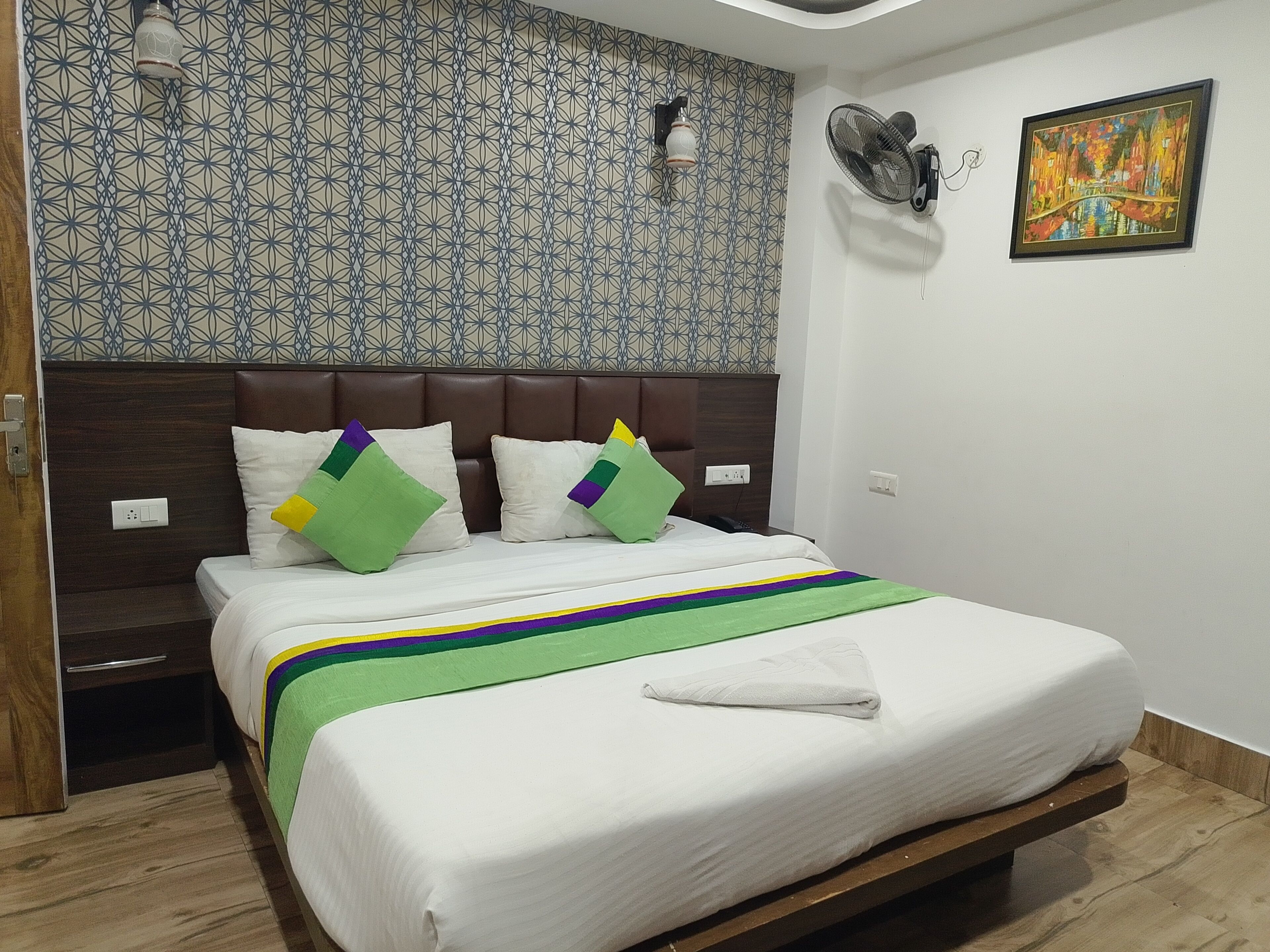 A hotel room with a bed, a headboard, a wall with a geometric pattern, a painting, and a fan.