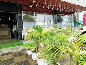 Entrada de hotel com varanda azulejada, plantas em vasos e toldo vermelho.