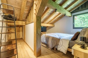 Ein Schlafzimmer im Loft-Stil mit Holzbalkendecke, Etagenbett, Schreibtisch mit Lampe und Fenster mit Blick auf Bäume.
