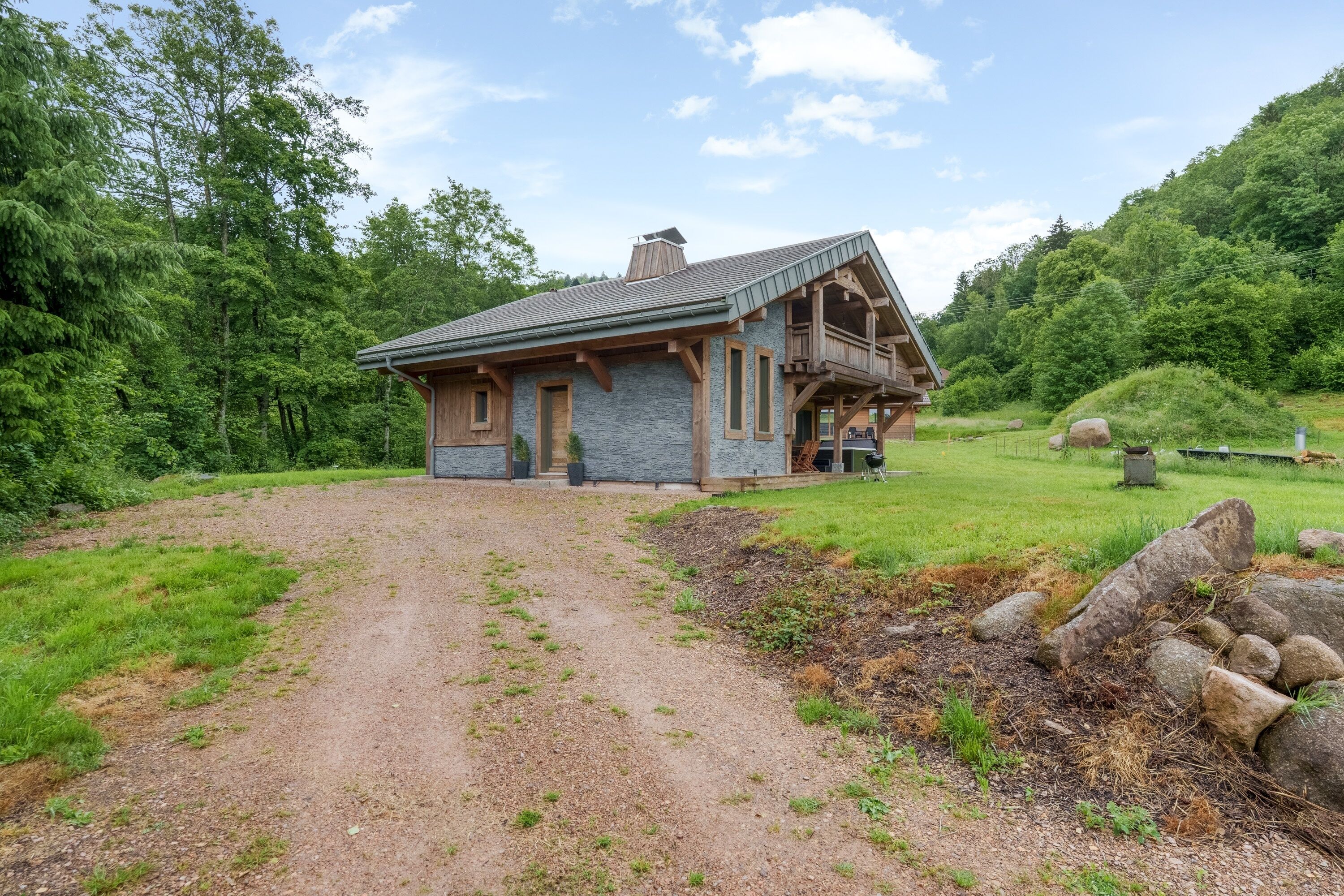 A rustic cabin with a stone exterior and a sloped roof, surrounded by a forested area and a gravel driveway.